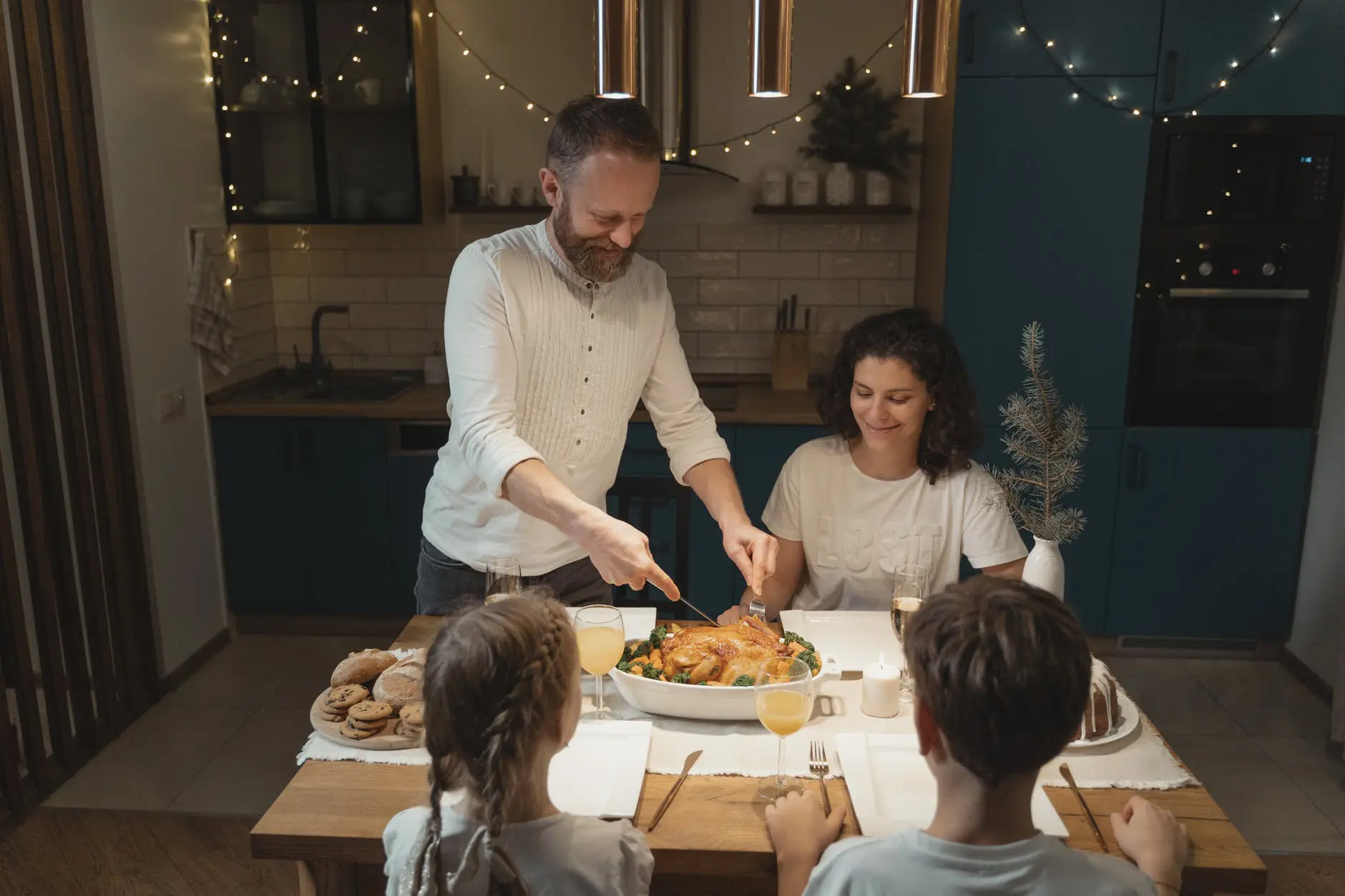 A man is carving a roast chicken at a festive dinner table with a woman and two children, evoking the warm hospitality one might experience on escorted tours of Ireland. The room, adorned with string lights, mirrors the enchanting ambiance of Irish celebrations.