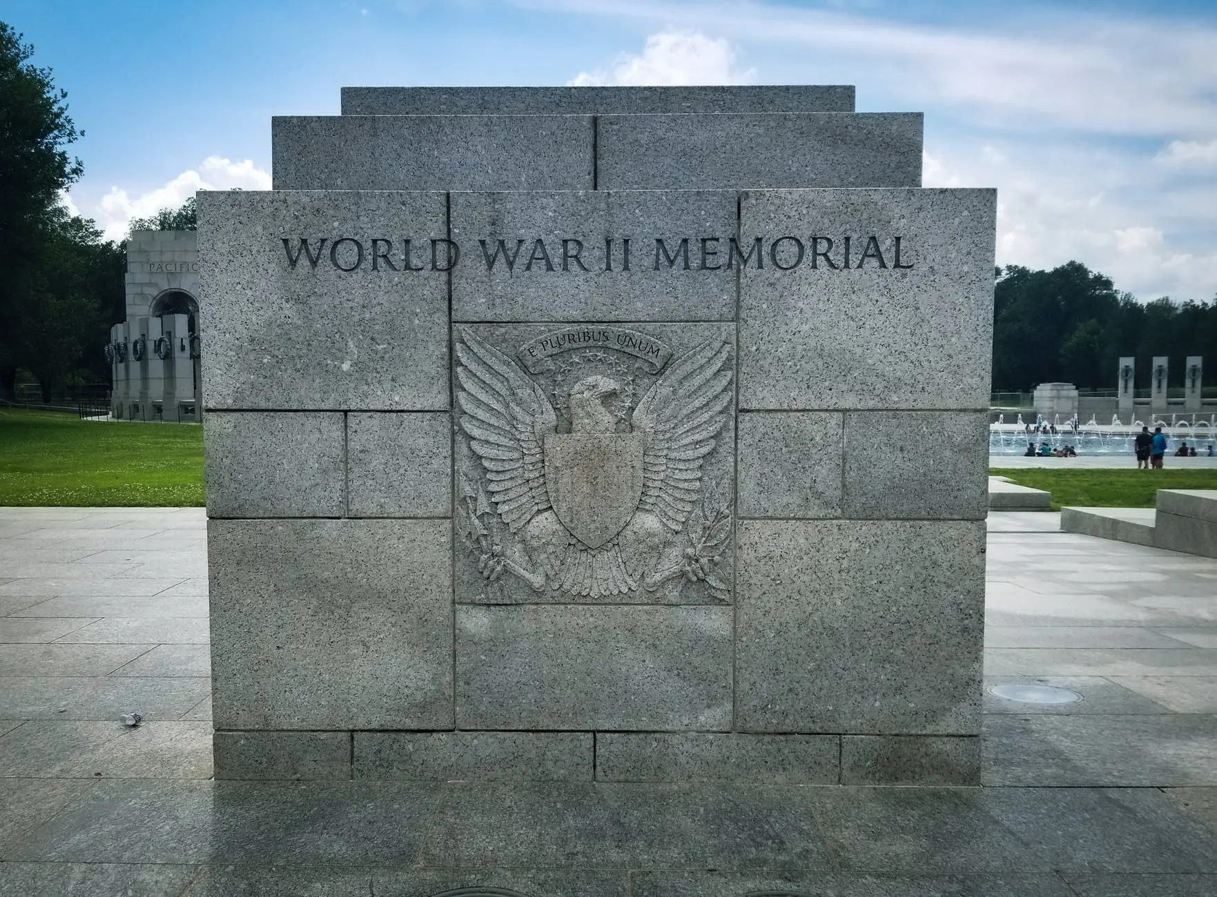 The stone entrance of the World War II Memorial, adorned with an eagle insignia, evokes solemn reflection. In the background, nestled alongside grass and other memorial structures, it stands as a poignant reminder for those on custom Ireland tours seeking historical insights across the globe.