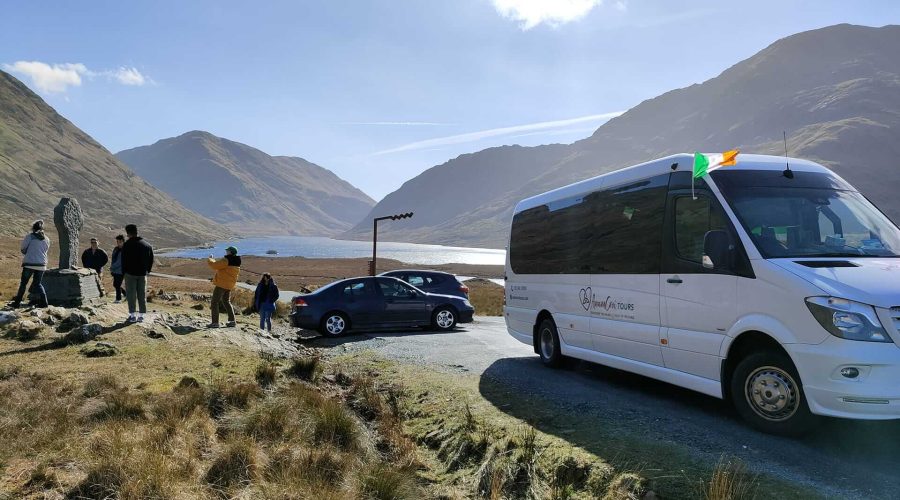 People stand by a parked tour bus and cars, overlooking a mountainous landscape with a lake. An Irish flag is attached to the bus, hinting at one of the premier Custom Ireland Tours—perfect for discovering hidden gems in comfort and style.